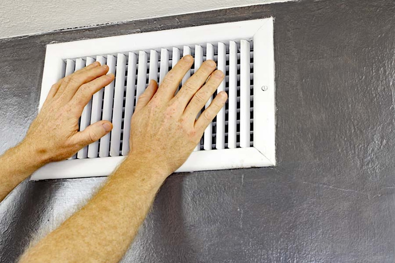 Why Is My Air Conditioner Blowing Hot Air? A pair of adult male hands feeling the flow of air coming out of an air vent on a wall near a ceiling. Man with hands in front of an air vent feeling for air flow.