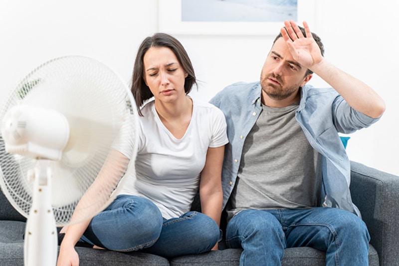 Why Is My AC Blowing Hot Air? Overheated couple sitting in front of a fan in their living room.