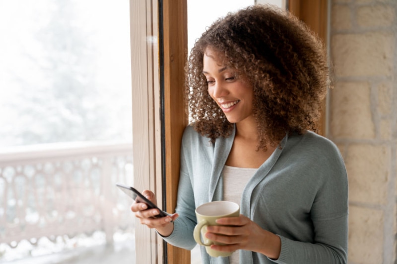 Why Choose a Variable-Speed Heat Pump? Image is a picture of woman texting in front of a window. Outside is snowy.