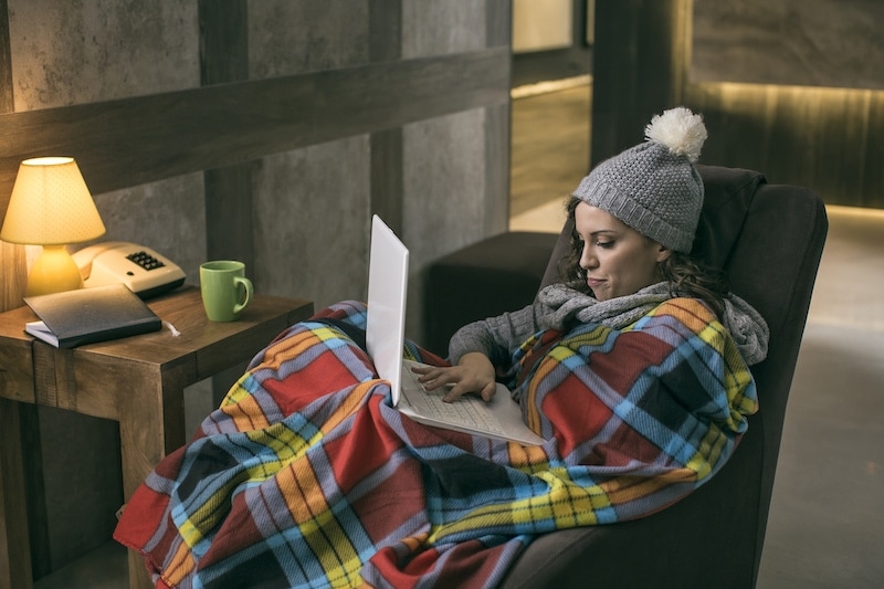 Young women wearing winter cap working on laptop wrapped in a blanket at home with no heating