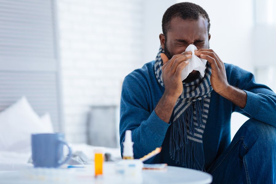 The Basics of Winter Indoor Air Quality. Image shows man wearing a scarf, sitting at table and blowing his nose into a tissue.